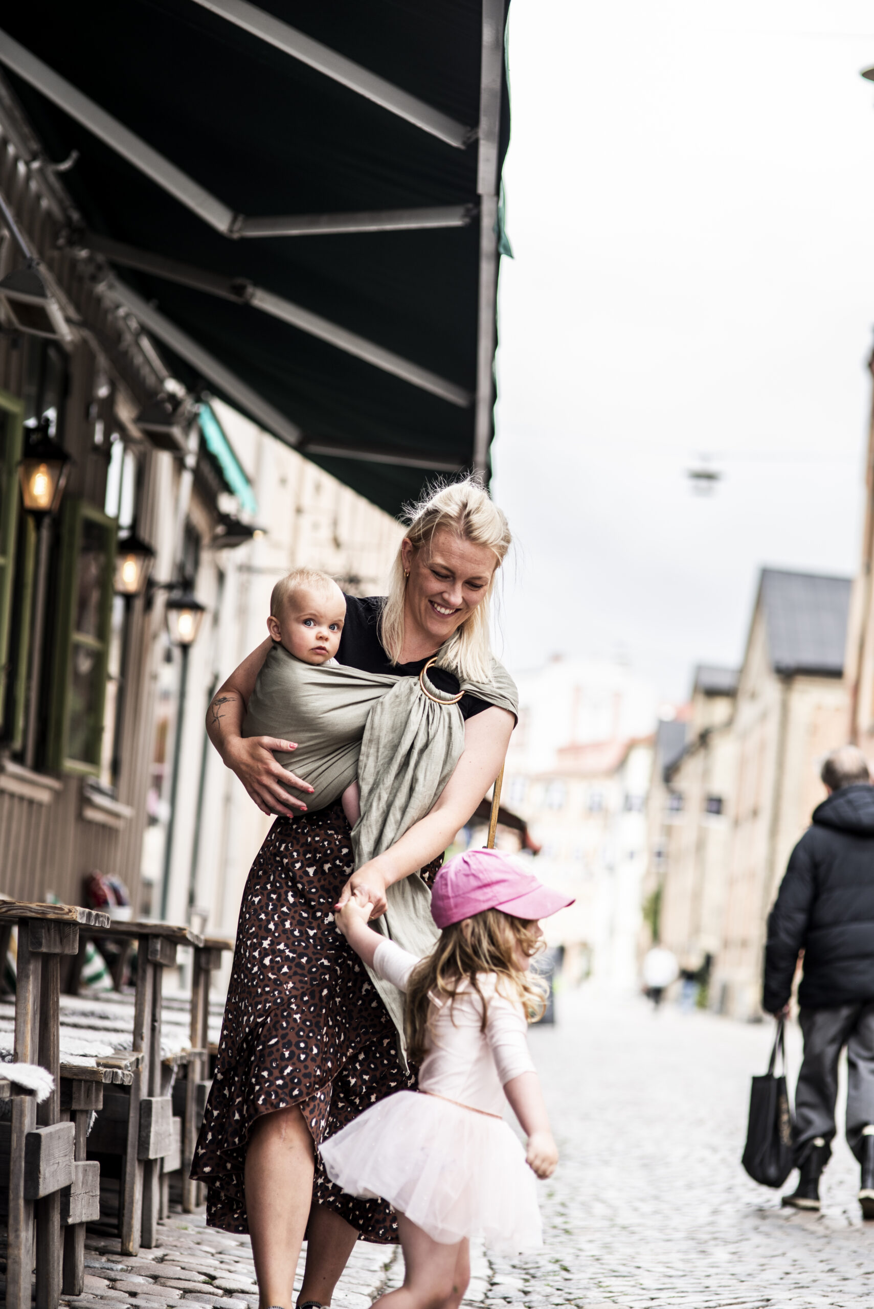 En mamma med ett barn i en bärsjal och ett annat barn som hon håller i handen. Bilden är tagen i stadsmiljö.